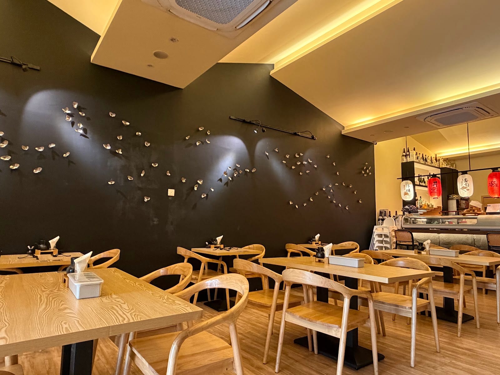 Don House dining room: a dark feature wall with a flock of small metal birds under spotlights, light wood tables and chairs, and the sushi counter with paper lanterns at the back
